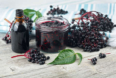Delicious homemade black elderberry syrup in glass jar and bunches of black elderberry with green leaves on wooden desk. Elderberry syrup immunity boosting for flu season.の写真素材