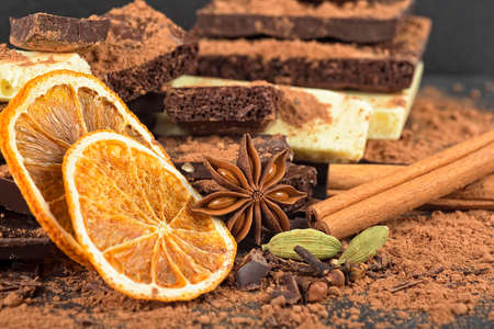 Broken chocolate pieces, cocoa powder, spices and dried oranges on a black background