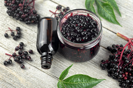 Homemade black elderberry syrup in glass jar and bunches of black elderberry with green leaves on wooden desk.の写真素材