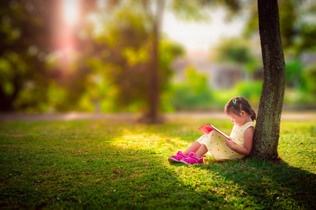 A little cute girl in a yellow dress reading a book sitting under the treeの写真素材
