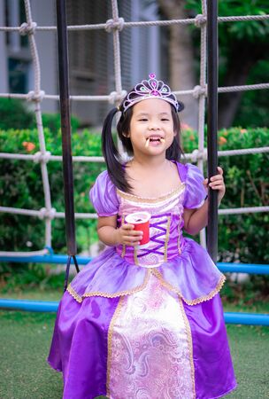 Portrait of cute smiling little girl in princess costume eating snack on swingの写真素材
