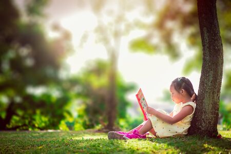 A little cute girl in a yellow dress reading a book sitting under the treeの写真素材