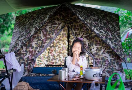 little asian girl sitting and eating breakfast in front of tent while going to camping.The concept of outdoor activities and adventures in natureの写真素材