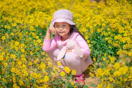 little asian girl in pink dress wear hat sitting in yellow flower gardenの写真素材