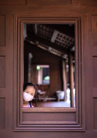 little asian girl wearing a mask against dusk and covid-19 near the windowの写真素材