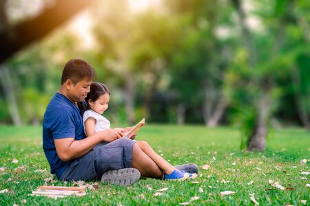 father sitting on grass with little daughter and reading book story in the parkの写真素材
