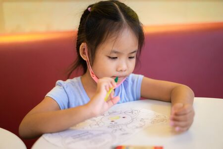 asian little girl sitting at table, drawing pictures with colored pencils in paper at home