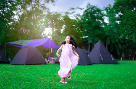 happy little asian girl in dress walking on the grass in front of tents while going camping with sunsetの写真素材