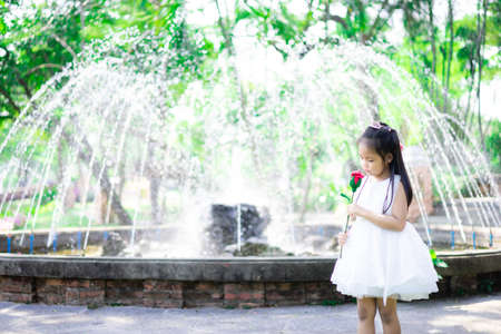 cute little asian girl in white dress holding a red rose in the parkの写真素材