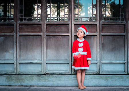 little asian girl in red Santa Claus costume with present box standing on wooden door  backgroundの写真素材