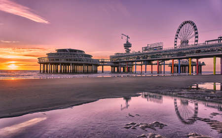 Scheveningen pier with ferris wheel and bungy jumping attractions reflecting in shallow water during low tide at sunsetのeditorial素材