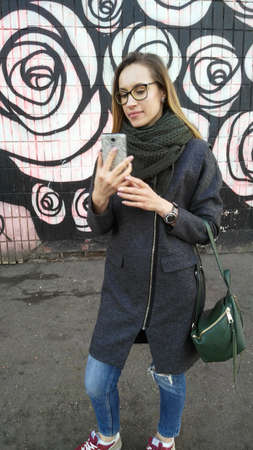A woman in glasses, a coat, jeans and a scarf takes a selfie against the background of a colored wallの写真素材