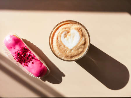 Pink eclair and a glass of cappuccino on a beige background in bright light with hard shadowsの写真素材