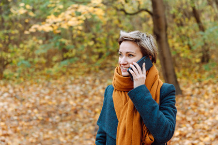 Short-haired blonde smiles as she talks on the phone in the fall outdoorsの写真素材