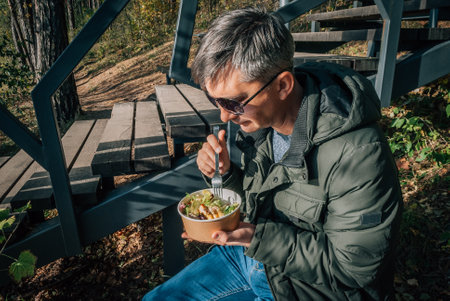 A man dines on a salad from a disposable paper bowl in the open air during a walk in nature in the fall.の写真素材