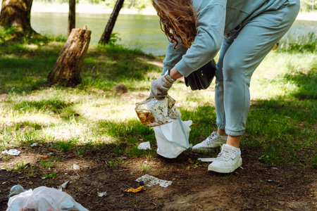 A woman cleans a picnic clearing in the forest near the river, collects bottles, plastic garbage and...