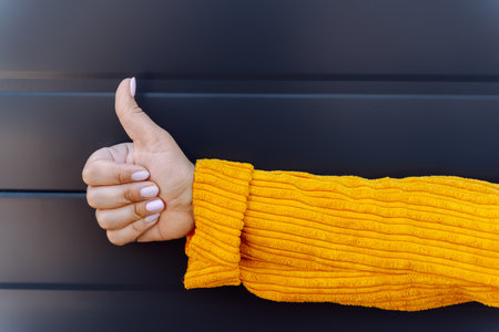 Person giving thumbs up in yellow sweater against dark striped background.の写真素材