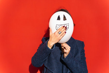 woman holding jack-o-lantern balloon and covering mouth in surprise against red background.