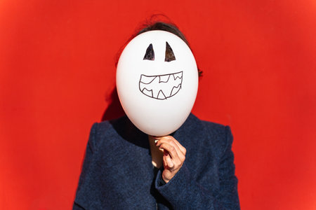 woman holding jack-o-lantern balloon in front of face against red background.の写真素材