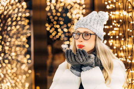 Young woman in knitted hat and down jacket blowing on gloved palms making a wish.の写真素材