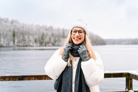 smiling woman adjusting her scarf on a winter day near a snowy lake.の写真素材