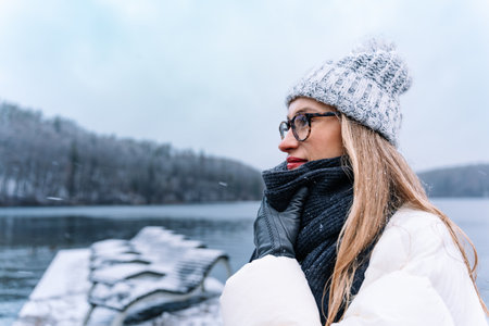 Woman wearing glasses and cozy winter attire, smiling, enjoying a snowy landscape by the lake.の写真素材