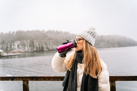 woman drinking from a travel mug by the lake on a snowy winter dayの写真素材