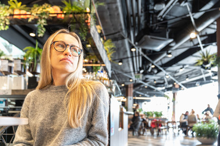 Stylish young woman with glasses and long blonde hair sitting in a modern cafe with urban decor, looking to the side. Relaxed atmosphere with natural light and industrial-style interiorの写真素材