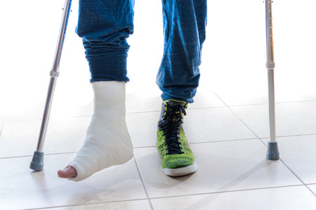 Young man with a broken ankle and a white cast on his leg following a basketball accident, walking on crutches and a high-top basketball shoe (isolated on white)の写真素材