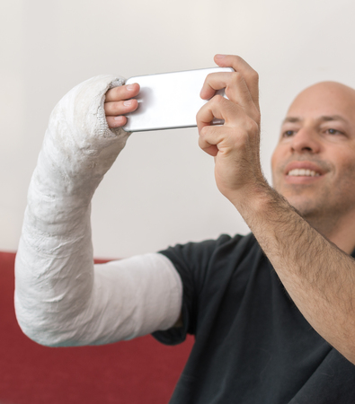Young man with an arm and elbow in a white plaster / fiberglass cast at home, talking a selfie on his smart phone while recovering from a broken armの写真素材
