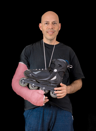 Young man sporting a bright red arm cast, holding an inline skate after a breaking his arm in a rollerblading accident, isolated on blackの写真素材