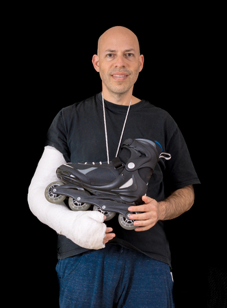 Young man sporting a bright white arm cast, holding an inline skate after a breaking his arm in a rollerblading accident, isolated on blackの写真素材