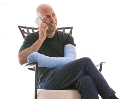 Young man with an arm and elbow in a blue plaster / fiberglass cast sitting on a modern couch, happily talking on his phone after having broken his armの写真素材