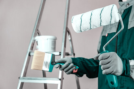 The plaster painter is ready to paint the wall. In the hands of a roller and brush. A stepladder and a bucket of paint in the backgroundの写真素材