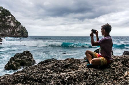 Young man sitting on the rock and making a photo of the wavesの写真素材