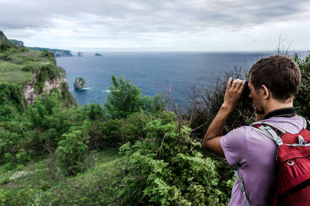 Young man with backpack on the hill making a photo of scenic landscapeの写真素材
