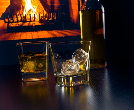 glasses of whiskey with ice cubes near whiskey bottle in front of the fireplace at nightの写真素材