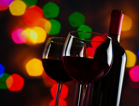 two red wine glasses near bottle against colorful bokeh lights background, festive and fun conceptの写真素材