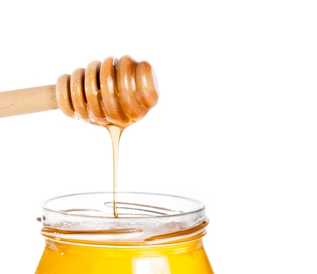 opened honey jar with wooden honey dipper on top pouring honey, isolated on white backgroundの写真素材