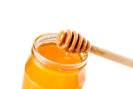 opened honey jar with wooden honey dipper on top pouring honey, isolated on white backgroundの写真素材