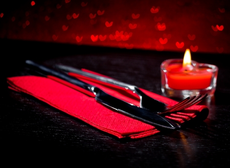 Valentine day table setting with knife, fork, red burning heart shaped candle on red hearts bokeh backgroundの写真素材