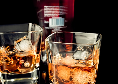 whiskey with ice in glass on black background near bottle, with reflectionの写真素材