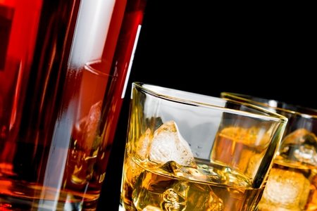 close-up of whiskey with ice in glasses near bottle on black background, with reflectionの写真素材