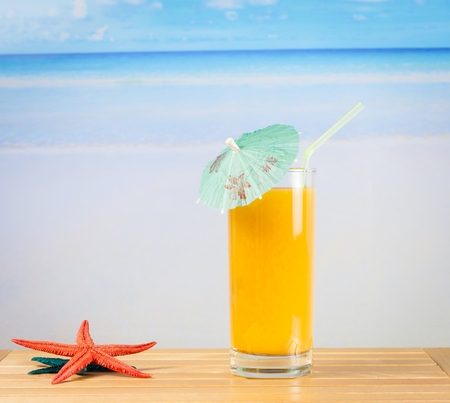 glass of orange juice on the sandy beach near starfish with sea in backgroundの写真素材