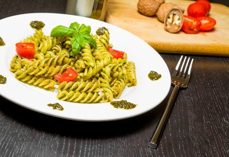 dish of pasta with pesto genovese sauce and vegetables on black wood table near fork, with ingredients, walnuts,  tomato, basil and olive oil in backgroundの写真素材