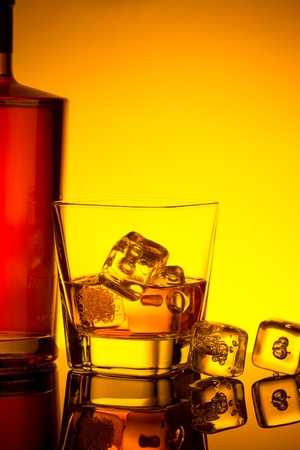 glass of whiskey with ice cubes near bottle on table with reflection, warm tint atmosphere, time of relax with whiskyの写真素材