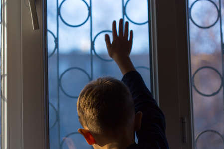 A boy stands and looks out the window at sunsetの写真素材