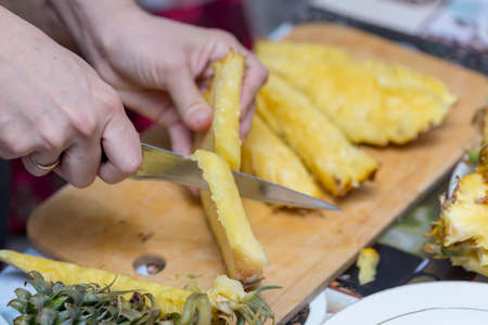 Hands of a man with a knife cut a juicy pineapple on a wooden board on the table around a dark background. Useful ingredientの写真素材