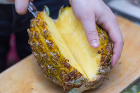 Hands of a man with a knife cut a juicy pineapple on a wooden board on the table around a dark background. Useful ingredientの写真素材