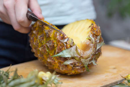 Hands of a man with a knife cut a juicy pineapple on a wooden board on the table around a dark background. Useful ingredientの写真素材